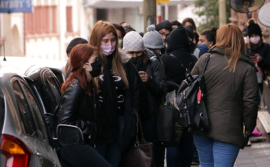Mujeres formando fila en una vereda, abrigadas y con mascarillas.