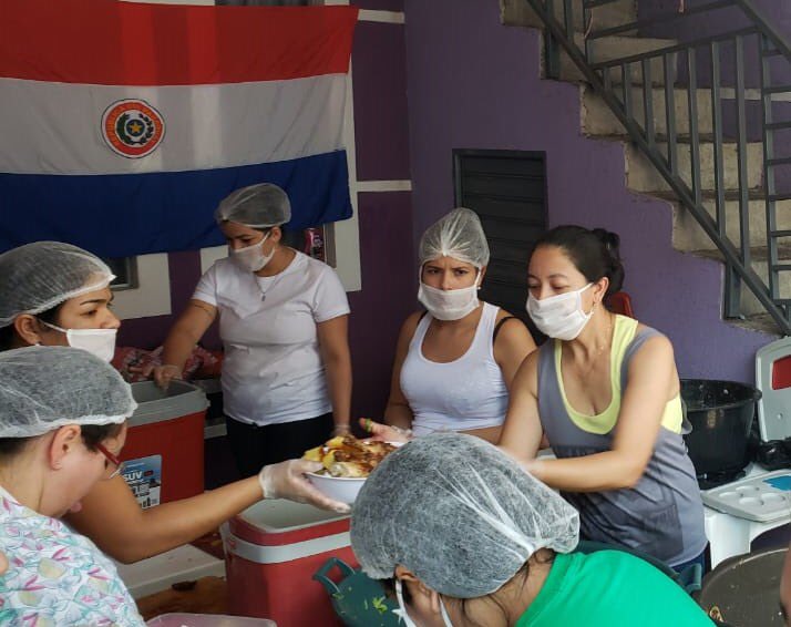 Mujeres brindando asistencia alimentaria a compatriotas. De fondo una bandera paraguaya.